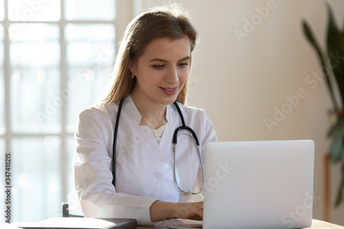 Successful young Caucasian female doctor in white medical uniform and stethoscope busy working on laptop in modern hospital, smiling woman nurse or GP typing on computer consult patient online