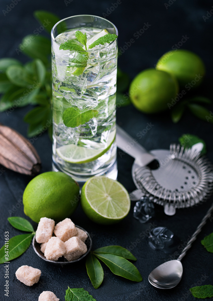 Fototapeta premium Highball glass of Mojito cocktail with ice cubes,mint and lime on black board with spoon and strainer and fresh limes with cane sugar and wooden squeezer and black shaker.