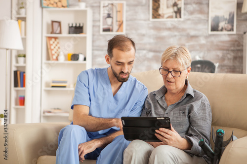 Slika na platnu Male bearded doctor in nursing home helping senior woman to use her tablet computer