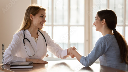 Smiling young woman GP shake hand greeting get acquainted with patient at consultation, happy Caucasian female doctor or physician handshake hospital clinic client closing deal at meeting