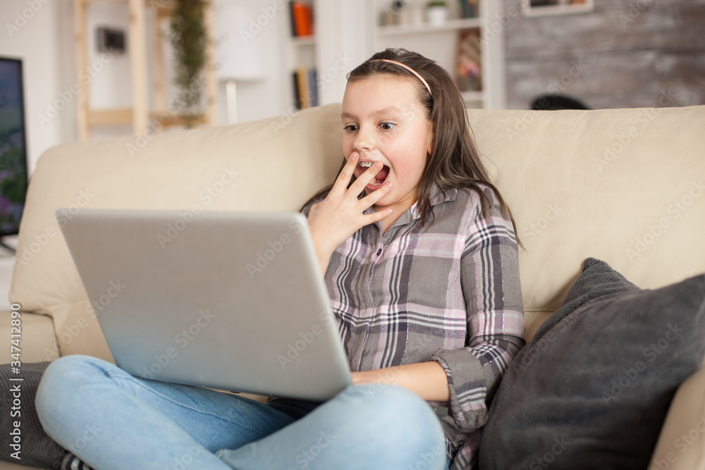 Fototapeta premium Excited little girl with braces while using her laptop sitting on the couch in living room.