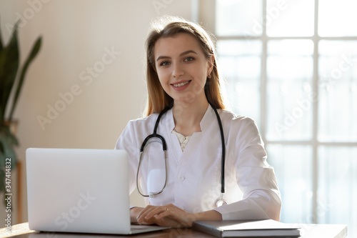 Portrait of successful young female doctor in white medical uniform and stethoscope sit at modern workplace with laptop, smiling millennial Caucasian woman nurse or GP work on computer in hospital