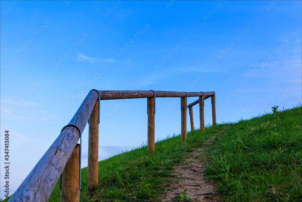 Corrimano, di legno, su sentiero di collina, prato verde e cielo sereno azzurro.