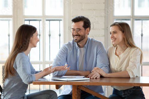 Happy young couple talk consult with female real estate agent or broker buying first house together, smiling millennial husband and wife discuss moving to new rent home with woman realtor at meeting