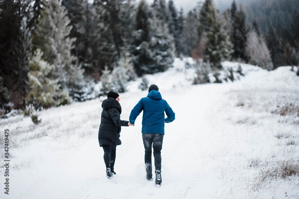 Beautiful young couple running in the mountains holding hands between Christmas trees in winter. Snow. Love of young couple. Free space for text is available.