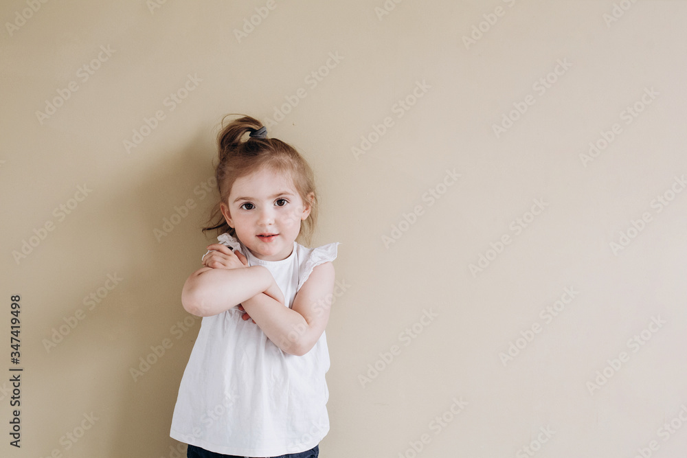 A happy little girl of European appearance stands on a beige background. Three year old baby in a white t-shirt. A happy little girl of European appearance stands on a beige background. Three year old baby in a white t-shirt.