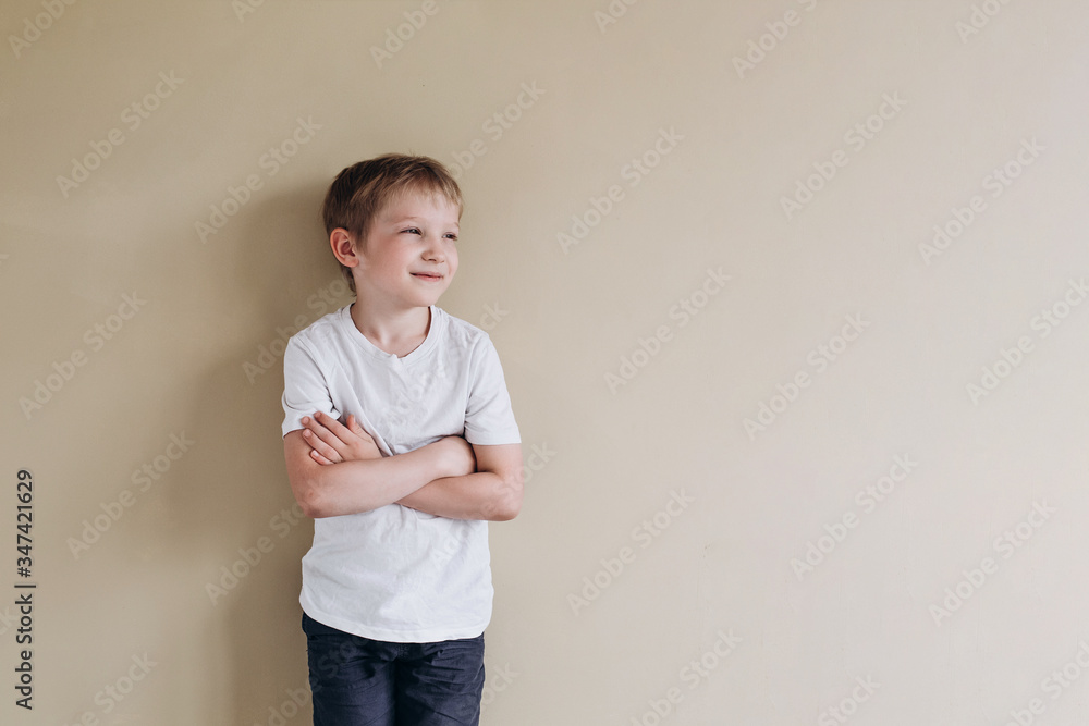 A happy boy of European appearance in a white T-shirt looks into the distance. Photo of a 7 years old baby on a beige background. A happy boy of European appearance in a white T-shirt looks into the distance. Photo of a 7 years old baby on a beige background.