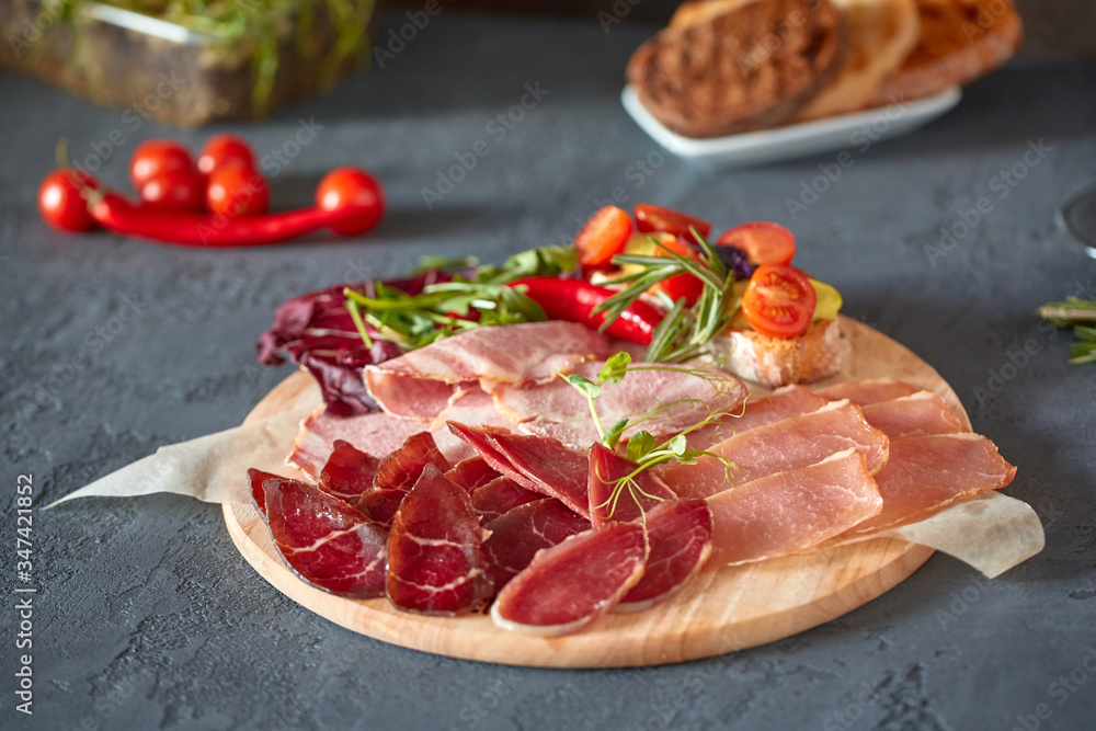 Antipasto on a wooden plate close-up. Cold smoked meat plate with sliced ham, prosciutto, and bacon. Decorated with bruschetta with fresh vegetables and herbs