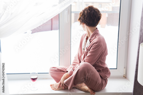 Beautiful woman wears pajamas with a glass of red wine sits on the windowsill near the beautiful window at home. Stay home. Quarantine and coronavirus covid-19 pandemic.