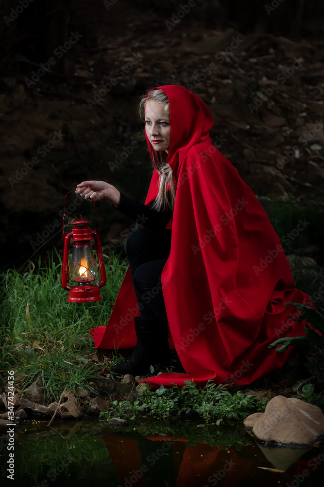 Attractive woman dressed a little red riding-hood cross stream in a dark with a lantern Photo | Adobe Stock