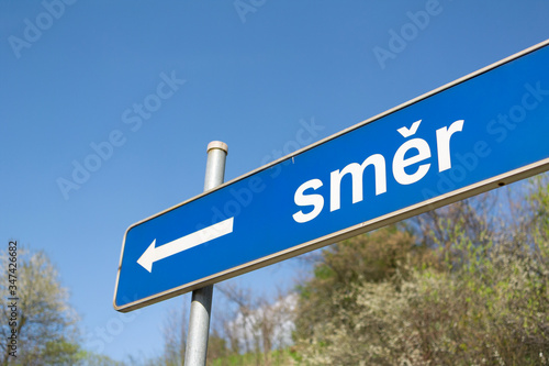 Traffic sign with symbol and pictogram of arrow and text (translation from Czech: Direction) - Navigation to left in Czechia / Czech Republic. Blue sky and green bush in the background. Shallow focus