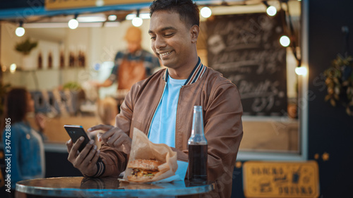 Handsome Young Indian Man is Using a Smartphone while Sitting at a Table in a Outdoors Street Food Cafe. He's Browsing the Internet or Social Media, Posting a Status Update. Man is Happy and Smiling.