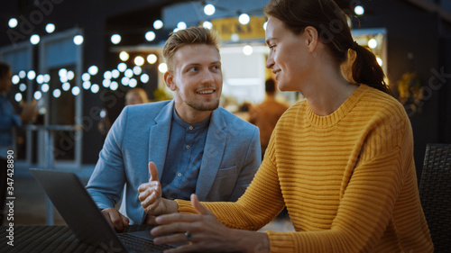 Beautiful Young Couple is Using a Laptop while Sitting at a Table in an Outdoors Street Food Cafe. They're Browsing Internet or Social Media, Posting a Status Update. They are Happy and Smile.