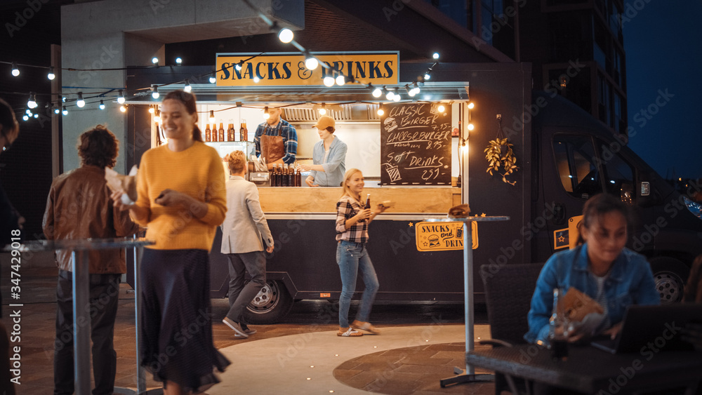Obraz premium Food Truck Employee Hands Out Beef Burgers, Fries and Cold Drinks to Happy Hipster Customers. People are Eating at Tables Outside. Commercial Truck Selling Street Food in a Modern Place Near the Sea.