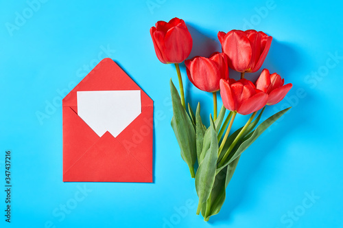 Bouquet of beautiful red tulips near envelope with blank paper sheet on blue background. Concept of celebrating valentine's, mother day or easter
