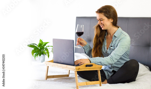 Happy Beautiful young woman teleworking at home in a bed with laptop and glass of red wine, lifestyle, copy space. Freelance and home office during coronavirus concept photo