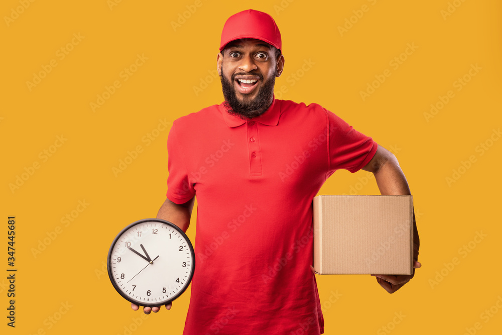 Joyful Courier Holding Cardboard Box And Clock Over Yellow Background Joyful Courier Holding Cardboard Box And Clock Over Yellow Background