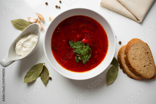 Beetroot soup in white bowl on a white table served with napkin, garlic, pepper, bread and saucy with sour cream. Traditional ukrainian, russian soup borscht with greens. Top view.