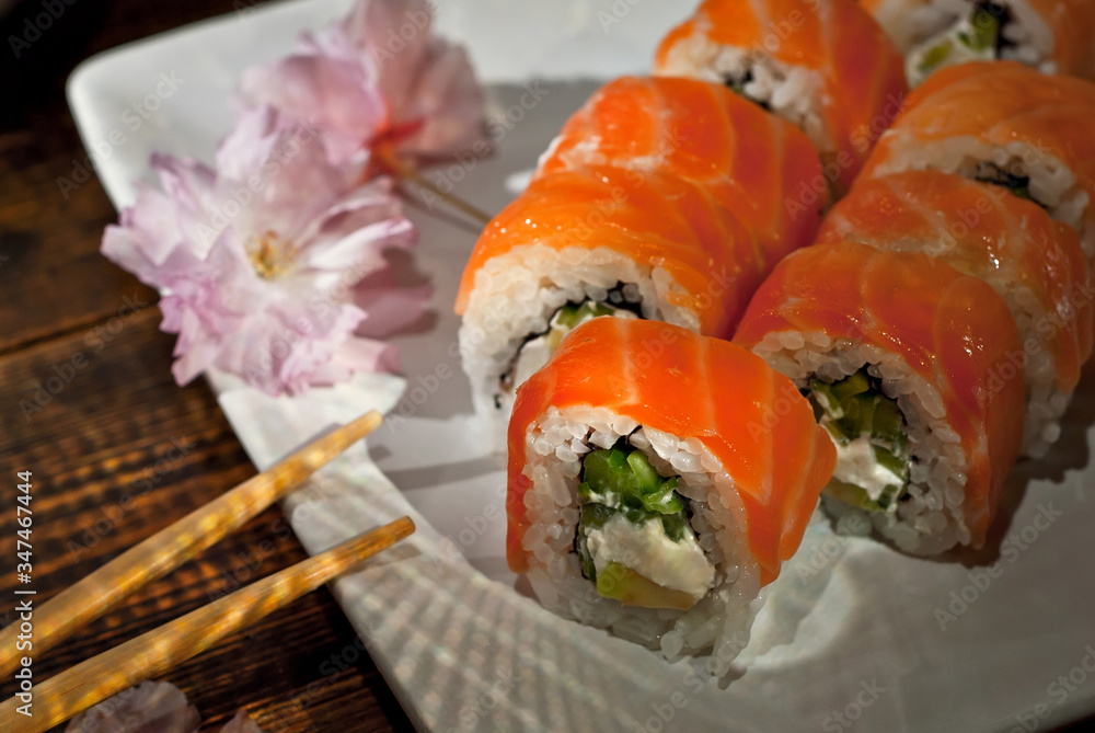Sushi on a white plate. Sushi roll with sauce and spices on a black background. Food on a wooden table with dark boards. Sakura color on a plate. Flower with rose petals. Sushi on a white plate. Sushi roll with sauce and spices on a black background. Food on a wooden table with dark boards. Sakura color on a plate. Flower with rose petals.
