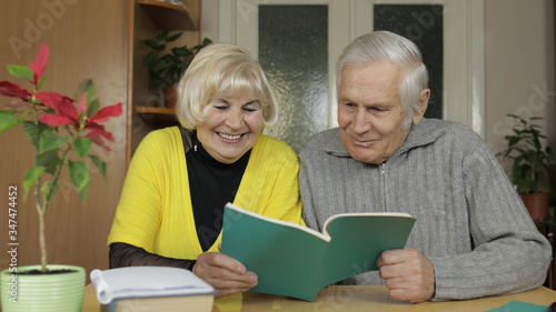 Happy old grandparents couple sit on table at home enjoy reading book together