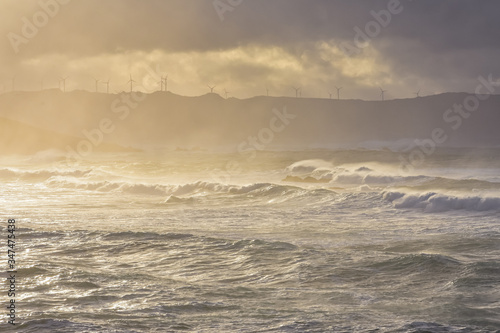 Big waves break on the coast, in the sunlit sea, passing through the clouds at sunset. Galicia, Spain. © dhvstockphoto