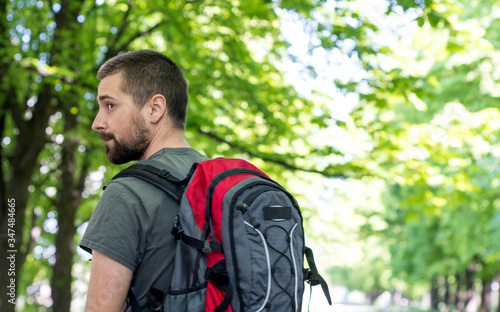 A young male tourist is walking in the park with a gray-red backpack and looking to the side. Back view. Nature travel concept with place for text.