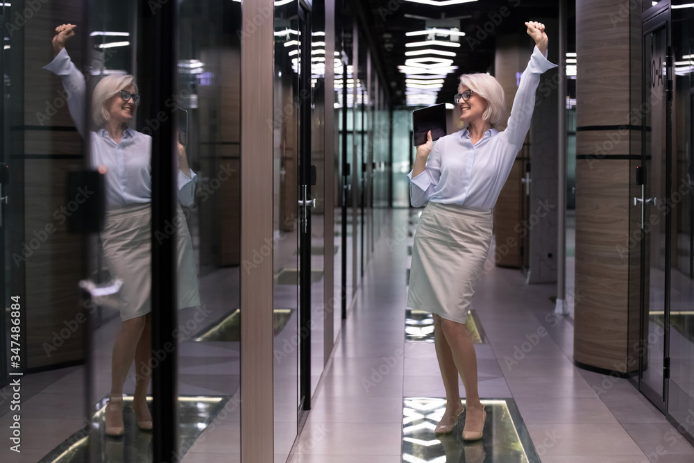 Elegant satisfied middle-aged businesslady company owner standing in office hallway dancing enjoy moment of business achievement and success, senior woman employee celebrate career advancement concept