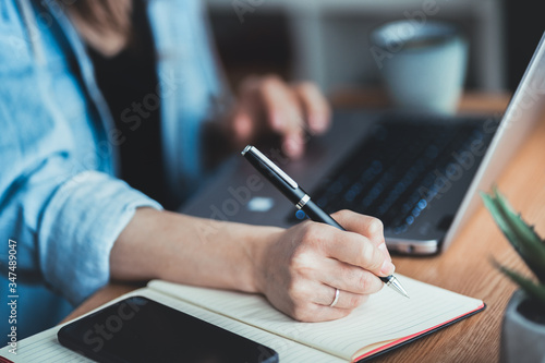 Young business woman sitting at desk, scrolling on laptop and taking notes in notebook. Close up of hand writing with pen