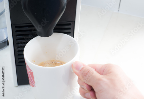 The man holds a white coffee mug with a right-hand waiting fresh coffee from the capsule coffee machine in the kitchen. photo