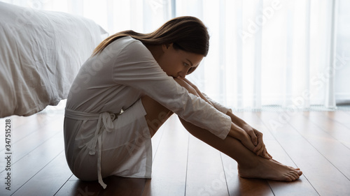 Sad young woman in white nightrobe sit barefoot on floor in bedroom leaned head to knees looking unhappy, suffers from loneliness, break up divorce with husband, hard decision about abortion concept photo
