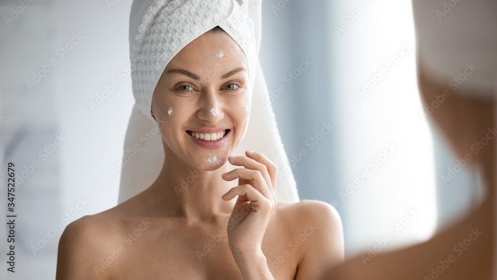 Over shoulder view female face reflected in mirror horizontal banner image, after shower with towel on head 30s woman apply moisturiser for dehydrated skin, self-care beauty treatment skincare concept Over shoulder view female face reflected in mirror horizontal banner image, after shower with towel on head 30s woman apply moisturiser for dehydrated skin, self-care beauty treatment skincare concept