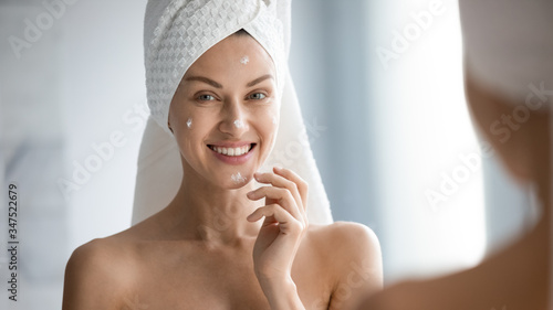 Over shoulder view female face reflected in mirror horizontal banner image, after shower with towel on head 30s woman apply moisturiser for dehydrated skin, self-care beauty treatment skincare concept