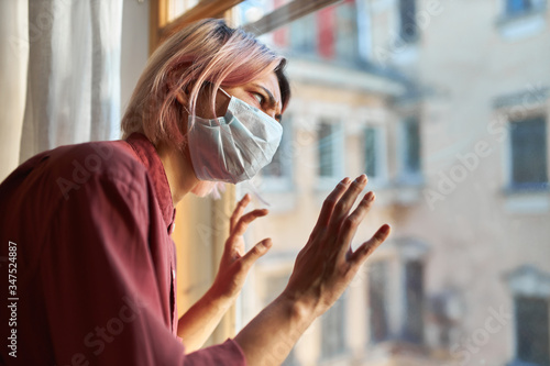 Young female patient with Covid-19 symptoms has to stay in hospital during quarantine, standing by window in disposable surgical mask, having stressed paranoid look, keeping hands on glass