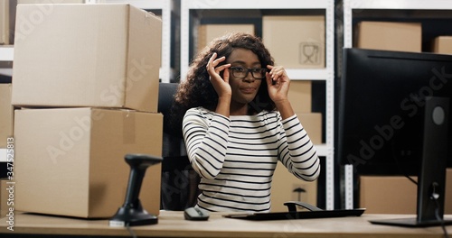 Portrait of happy young African American woman sitting at desk in postal office store working at computer.