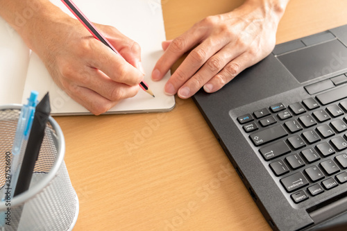 laptop, coffee cup, notebook on the wooden table. hands of businessman typing laptop keyboard at afternoon. a adult man in casual clothing is working on the chair for work from home.