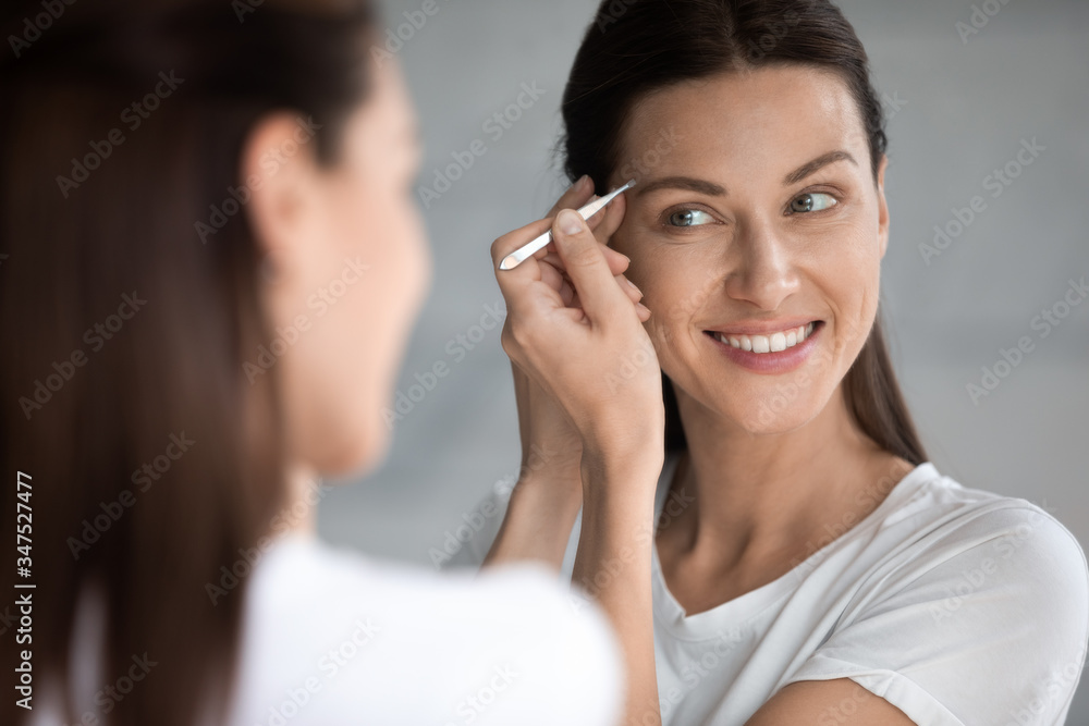 Over shoulder view female face reflected in mirror, woman hold metal tweezers plucks eyebrows corrects shape remove grow hairs feels satisfied, personal grooming beauty salon at home self-care concept Over shoulder view female face reflected in mirror, woman hold metal tweezers plucks eyebrows corrects shape remove grow hairs feels satisfied, personal grooming beauty salon at home self-care concept