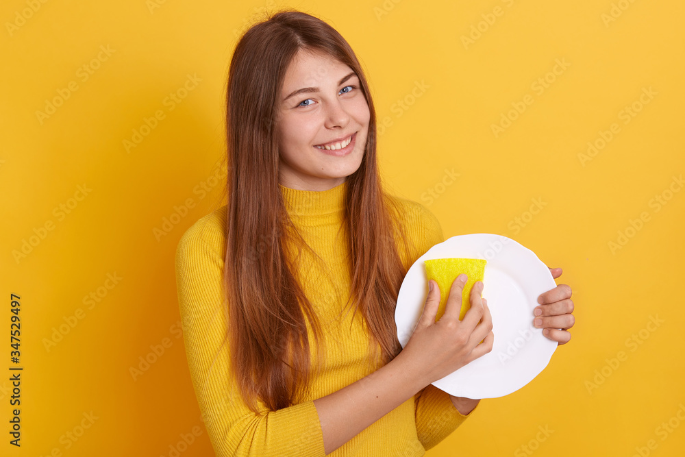 Happy smiling woman posing isolated over yellow studio background, looking directly at camera, washing plate with sponge, dresses casual clothing, being in good mood, washes dish after breakfast.
