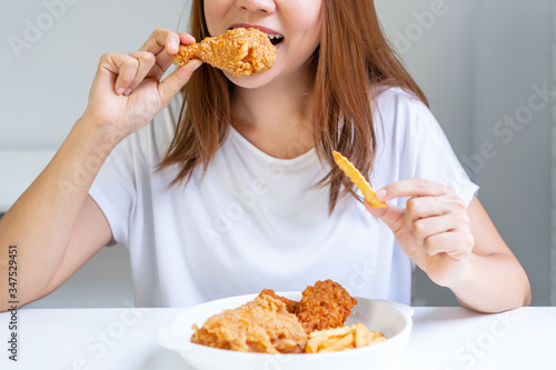 Close up portrait of a satisfied pretty Asian woman eating fried chicken and french fries isolated over white background. Unhealthy food concept, close up