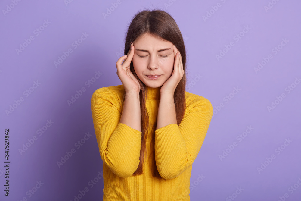 Studio shot of female wearing yellow casual shirt, keeping hands on her temples, keeps eyes closed, suffering from terrible pain, stands against lilac wall. Health care, people, medicine concept. Studio shot of female wearing yellow casual shirt, keeping hands on her temples, keeps eyes closed, suffering from terrible pain, stands against lilac wall. Health care, people, medicine concept.