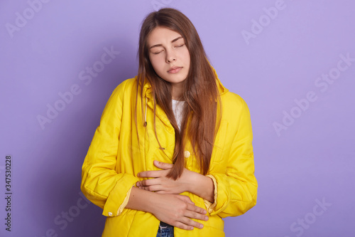 Studio shot of suffering from abdominal pain girl, lady dresses yellow jacket keeping hands on belly, keeps eyes closed, posing against lilac wall. People, health care, emotions, period concept.