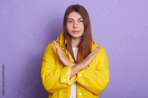 Indoor shot of serious or sad woman posing isolated over lilac studio background, lady wearing yellow jacket, crossing hands, female showing stop gesture. Body language, people, lifestyle concept. © sementsova321