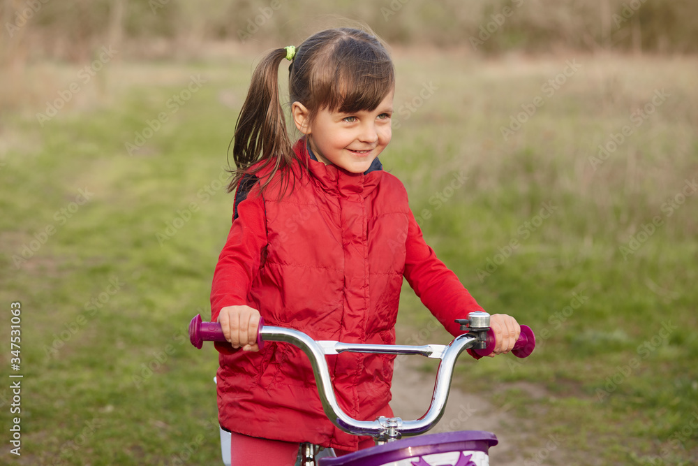 Obraz premium Outside picture of cheerful joyful little girl with pony tails, riding bicycle alone, having fun, breathing fresh air, being fond of nature, playing game, having delighted facial expression.