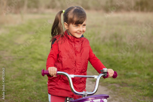 Outside picture of cheerful joyful little girl with pony tails, riding bicycle alone, having fun, breathing fresh air, being fond of nature, playing game, having delighted facial expression.