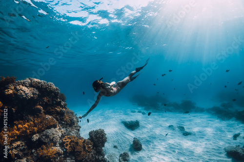 Woman free diver with fins glides over sandy bottom near corals underwater in blue sea