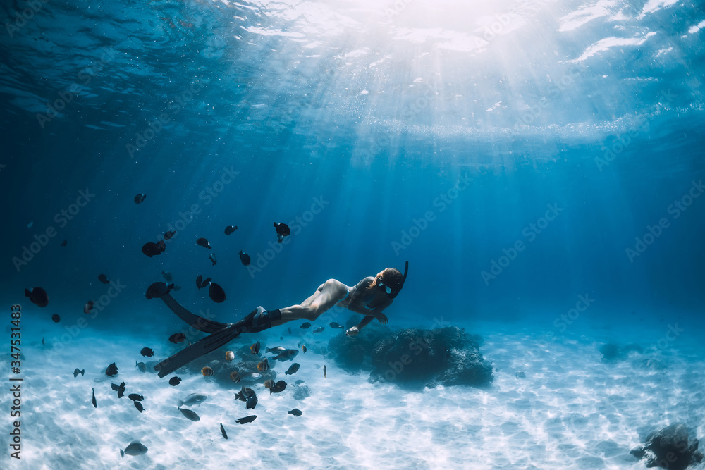 Free diver girl with fins glides over sandy bottom with tropical fishes in blue sea