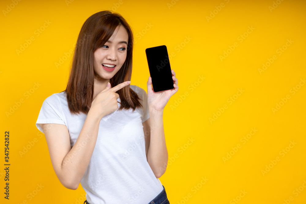 Young elegant Asian woman smiling and pointing to smartphone isolated on yellow background Young elegant Asian woman smiling and pointing to smartphone isolated on yellow background