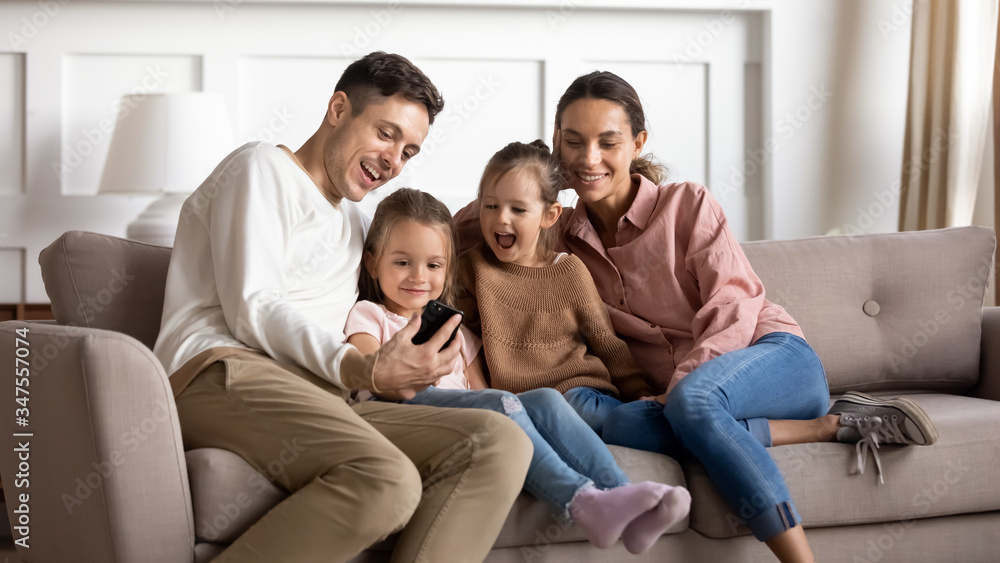 Happy parents with two little daughters using phone together, sitting on cozy sofa in living room, smiling father holding smartphone, preschool girls watching video online, spending weekend at home Happy parents with two little daughters using phone together, sitting on cozy sofa in living room, smiling father holding smartphone, preschool girls watching video online, spending weekend at home