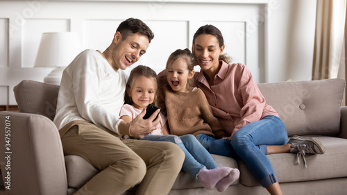 Happy parents with two little daughters using phone together, sitting on cozy sofa in living room, smiling father holding smartphone, preschool girls watching video online, spending weekend at home