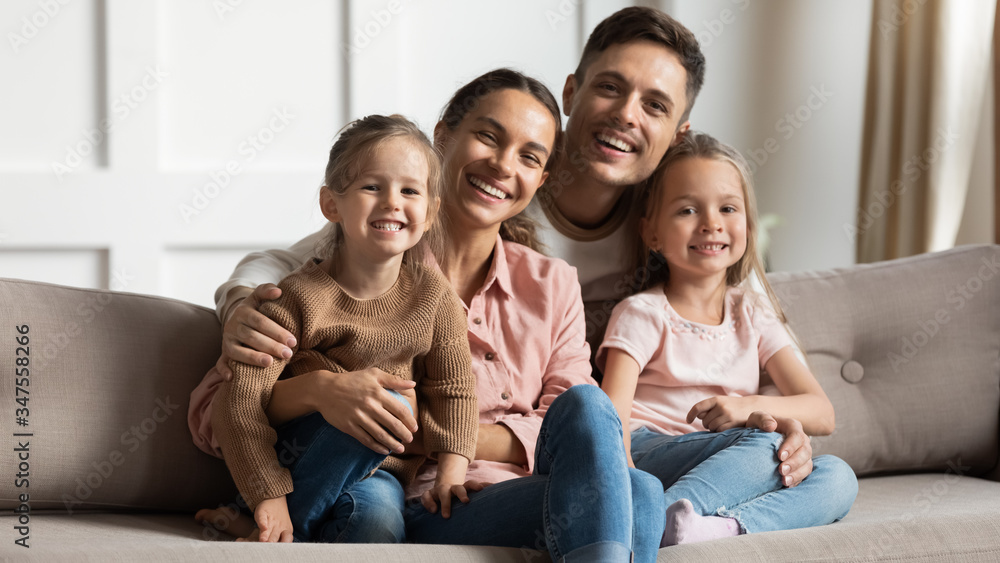 Portrait of smiling mother and father with two little daughters sitting on couch, looking at camera, happy parents hugging two preschool girls sisters, large family posing for photo at home Portrait of smiling mother and father with two little daughters sitting on couch, looking at camera, happy parents hugging two preschool girls sisters, large family posing for photo at home