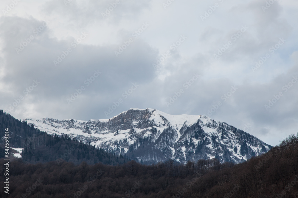A snowy mountain range in a cloudy sky and dark forest. Winter cloud landscape of mountains. Mountains Sochi, Russia